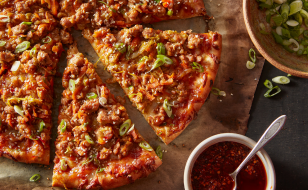 Sliced pizza topped with ground pork and assorted vegetables with bowl of chili sauce beside it.