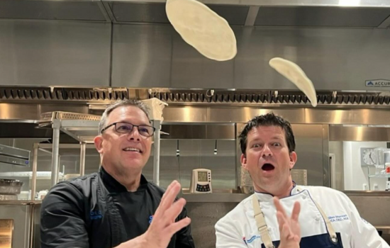 Two male chefs playfully tossing pizza rounds in the air.